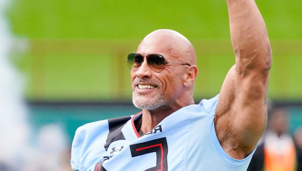 dwayne johnson wearing sunglasses and a light blue sports jersey raises his arm in celebration on a football field, smiling confidently with a blurred crowd in the background.