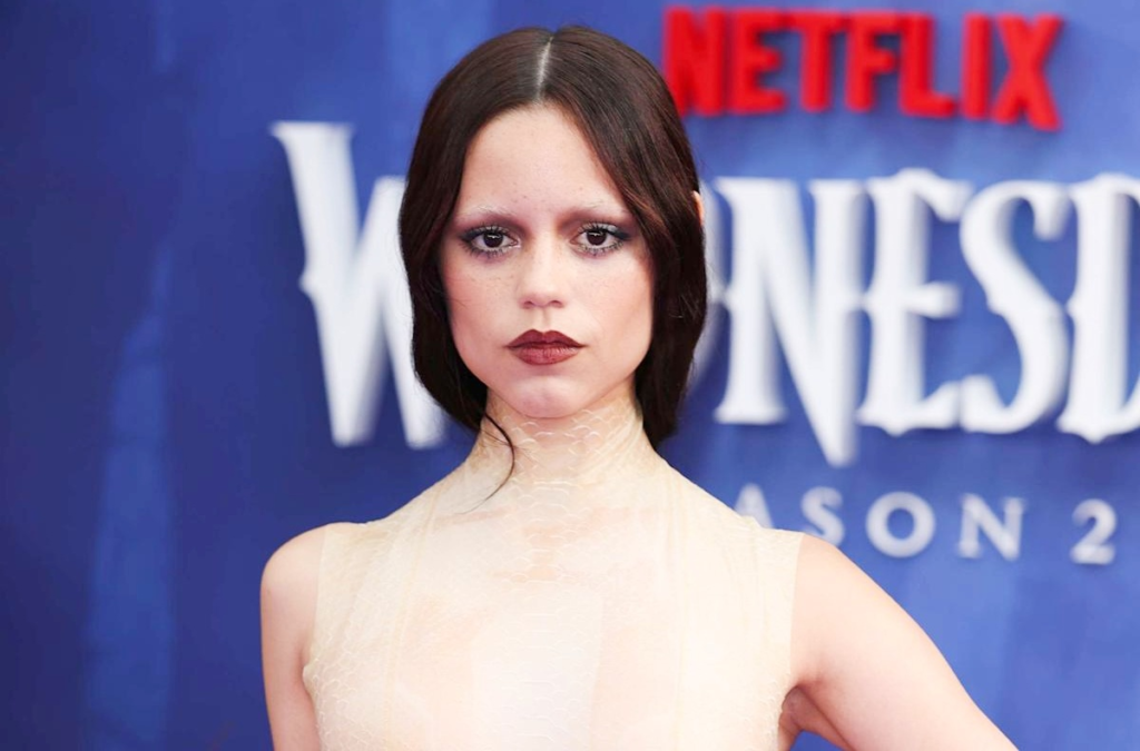 A woman in a pale textured gown posing on a blue Netflix event backdrop, captured with dramatic makeup and a serious expression, suitable for articles discussing Jenna Ortega net worth and her rising celebrity status.
