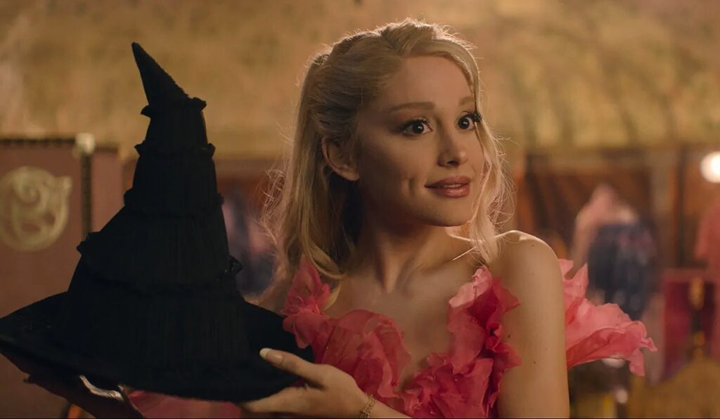 Ariana Grande wearing a pink ruffled dress, holding a black witch hat while smiling in a warmly lit indoor setting.
