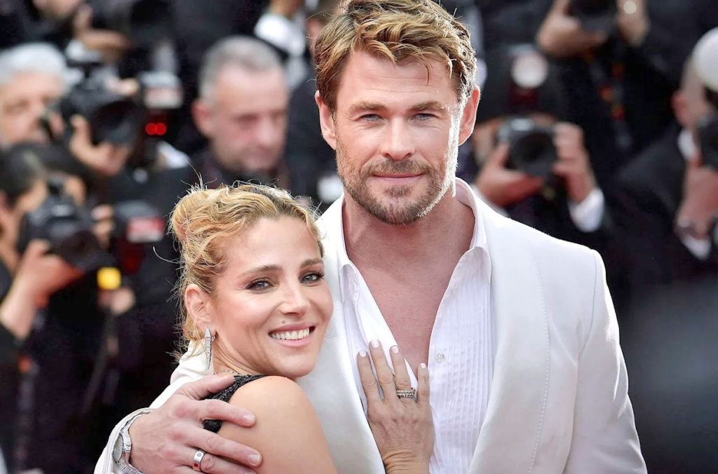 Chris Hemsworth, the Australian actor famous for playing Thor, embraces his beautiful wife Spanish actress and model Elsa Pataky on the red carpet. Both radiate elegance in coordinated white ensembles while surrounded by flashing cameras at a major 2025 film premiere or gala event – ultimate Hollywood couple goals.