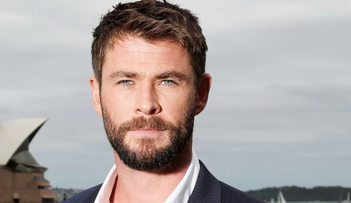A man with short brown hair and a well-groomed beard wearing a dark suit, posing outdoors with a confident look, showcasing a style inspired by Chris Hemsworth.