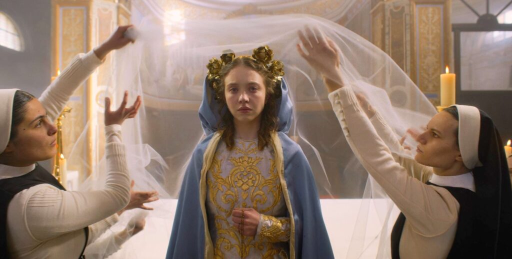 Two women dressed as attendants adjust a long flowing veil behind a woman in ornate medieval-style clothing inside a grand, golden-lit hall. Focus keyword: Sydney Sweeney.