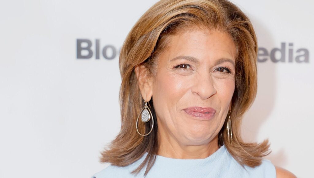Hoda Kotb with shoulder-length hair wearing a light outfit, standing in front of a media backdrop and smiling slightly