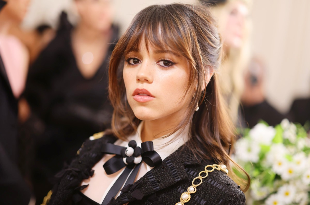 A close-up photo of a young woman with soft makeup, layered brown hair, and a black-and-white outfit at a formal event, used for articles discussing Jenna Ortega net worth and celebrity income profiles.