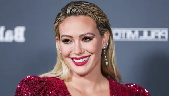 Red carpet portrait used in content related to Hilary Duff, showing a smiling woman in a sequined red dress with glamorous makeup and earrings.”