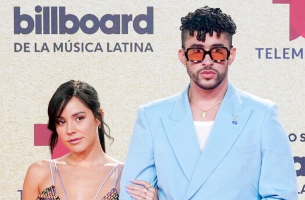 Bas Bunny posing on the red carpet at a Latin music event, wearing a light blue suit and tinted sunglasses, standing beside a woman in a stylish dress.”