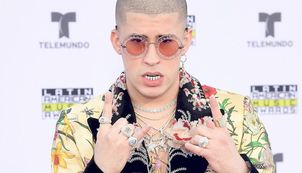 A person wearing a bold, Bad Bunny–style outfit featuring oversized round sunglasses, multiple rings, layered chains, and a patterned floral jacket while posing on a red carpet backdrop.