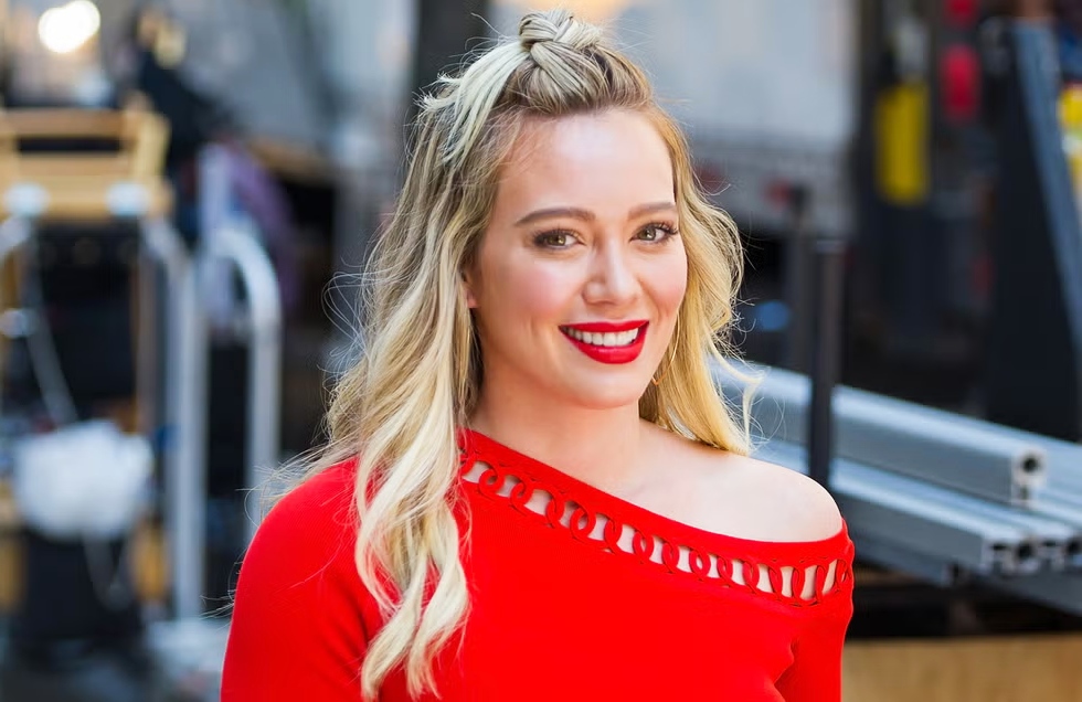 “A promotional-style photo used in content related to Hilary Duff, showing a smiling woman in a bright red off-the-shoulder top with wavy blonde hair, standing outdoors with filming equipment blurred in the background.