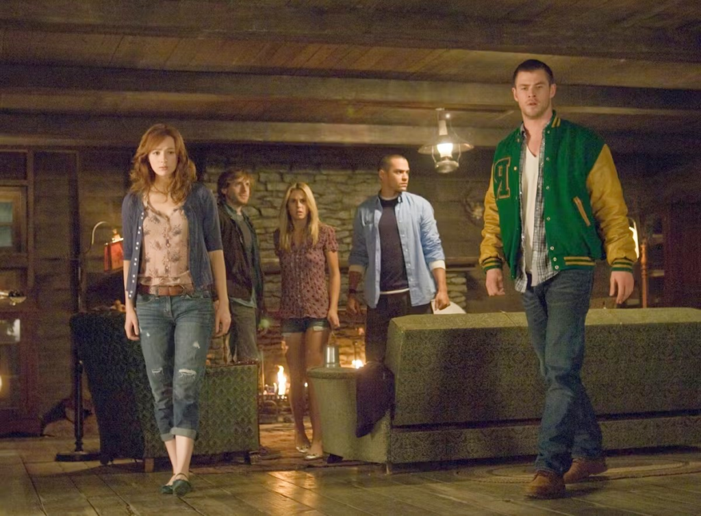 Young Chris Hemsworth, wearing a green-and-yellow letterman jacket, leads the group through the eerie wooden cabin living room in the 2011 horror-comedy classic The Cabin in the Woods. Co-stars Kristen Connolly, Fran Kranz, Anna Hutchison, and Jesse Williams follow behind in this iconic scene that launched Hemsworth’s Hollywood career just before Thor.