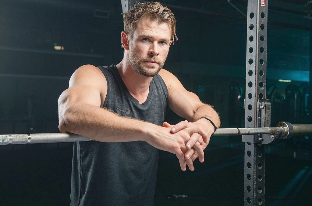 Australian actor Chris Hemsworth, best known as Thor in the Marvel Cinematic Universe, displays his jacked arms and muscular build during an intense gym workout session, leaning confidently on a loaded barbell in a sleeveless black Nike tank top – trending fitness inspiration 2025