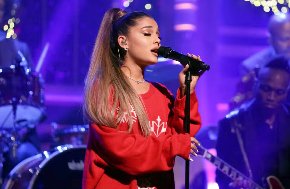 Ariana Grande performing live on stage wearing a red oversized sweater, singing into a microphone with her signature high ponytail under purple concert lights