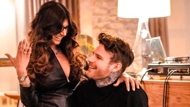  Mia Khalifa sharing a joyful moment indoors, smiling and leaning close to each other near a wooden table with a record player and a lamp in the background.