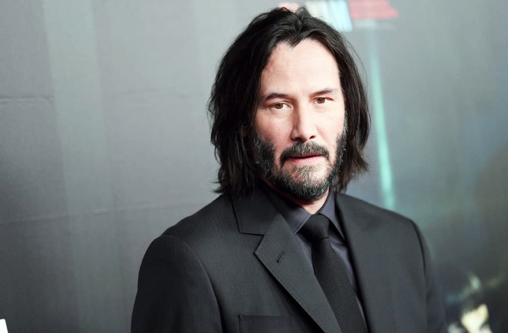 Keanu Reeves with shoulder-length dark hair and a beard is wearing a black suit, black shirt, and black tie. He is facing the camera with a neutral expression, and the background is plain and softly lit.
