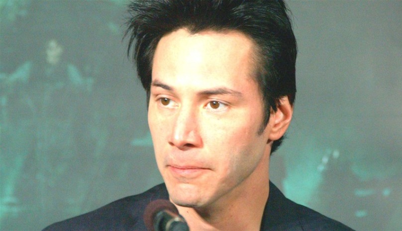 A man identified as Keanu Reeves is shown in the image with short, dark hair and a clean-shaven face. He is wearing a dark-colored shirt and looking slightly to the side with a calm expression. The background is blurred in shades of green and gray.