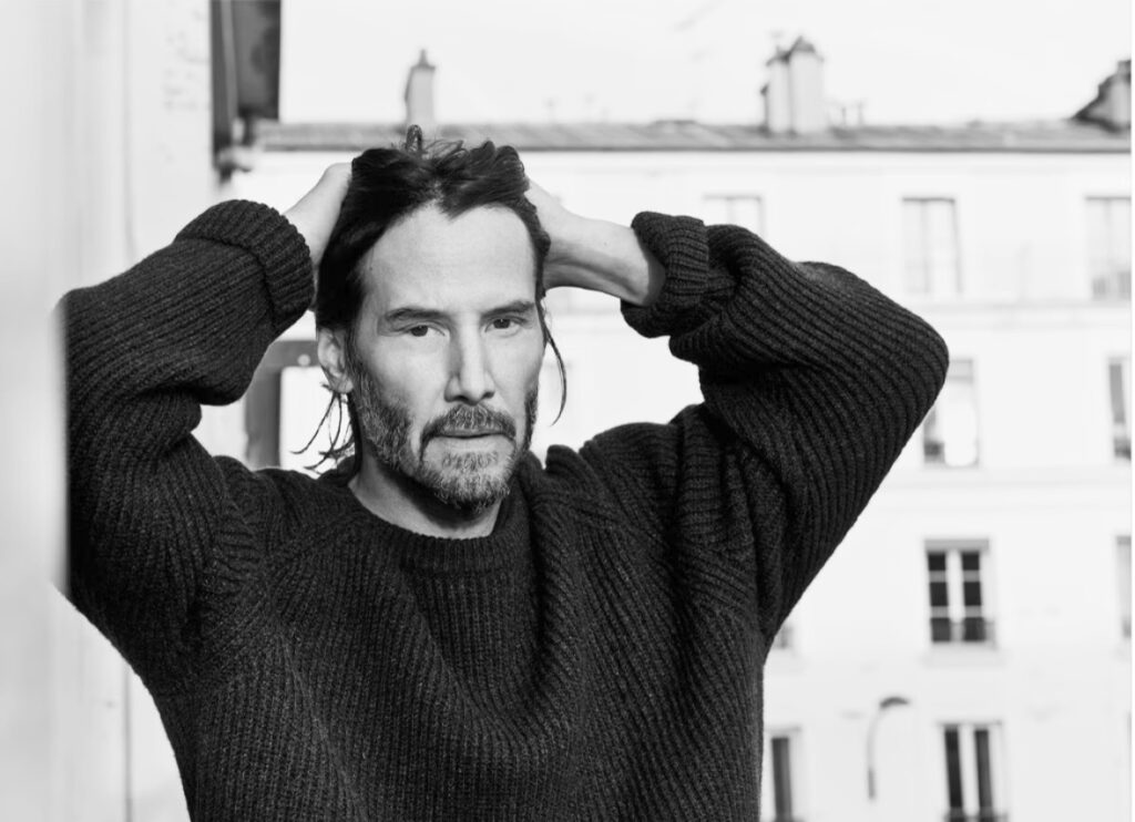 The image shows a man named Keanu Reeves wearing a dark, ribbed sweater. He has medium-length hair and a beard, and he is posing with both hands behind his head. The background appears to be indoors with a softly lit, neutral setting.