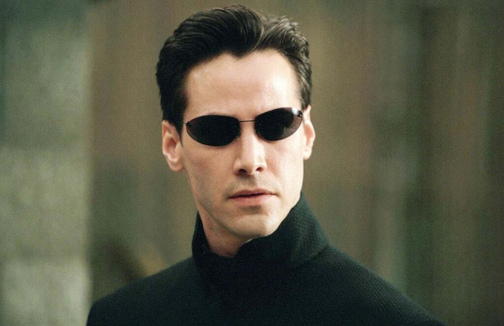 The image shows a man named Keanu Reeves wearing a black outfit and dark sunglasses. He has a serious expression on his face, and the background is blurred with greenish-gray tones.