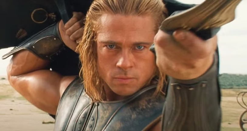 brad pitt with long blond hair and intense expression is dressed in ancient-style armor, holding a sword pointed forward. He appears to be in a battle scene, with a background that looks like an old stone or wooden structure.