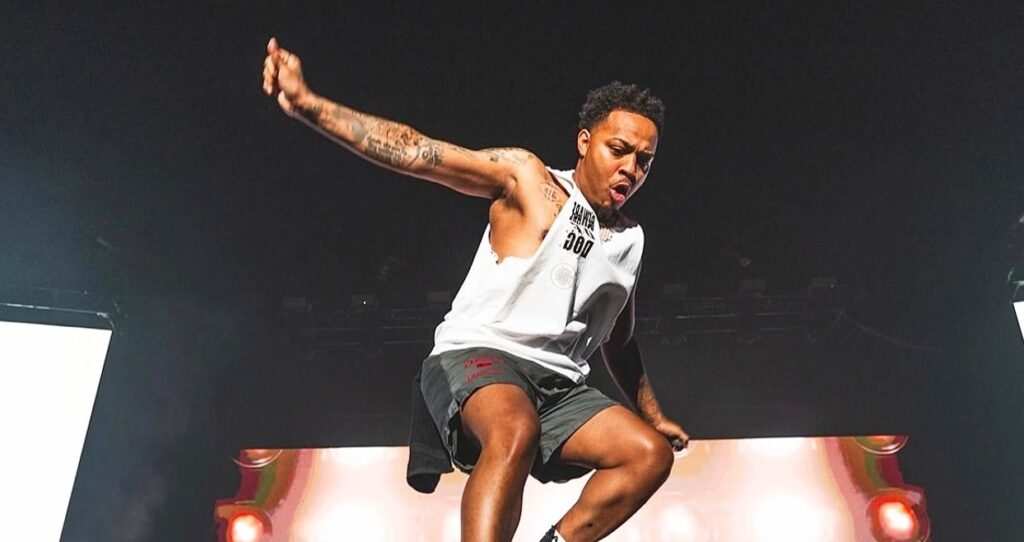Bow Wow performing energetically on stage, wearing a white sleeveless shirt and gray shorts. He is captured mid-air in a dynamic pose with one arm raised and a focused expression, under bright stage lighting against a dark background.