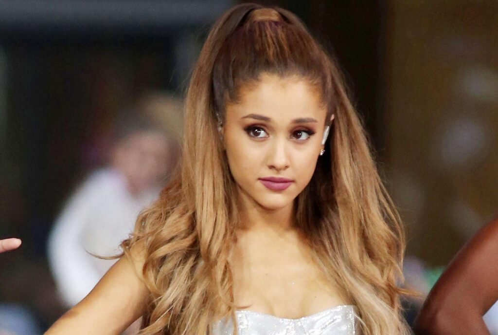 Ariana Grande with long wavy light brown hair, wearing a silver outfit and natural glam makeup, posing gracefully at an event.