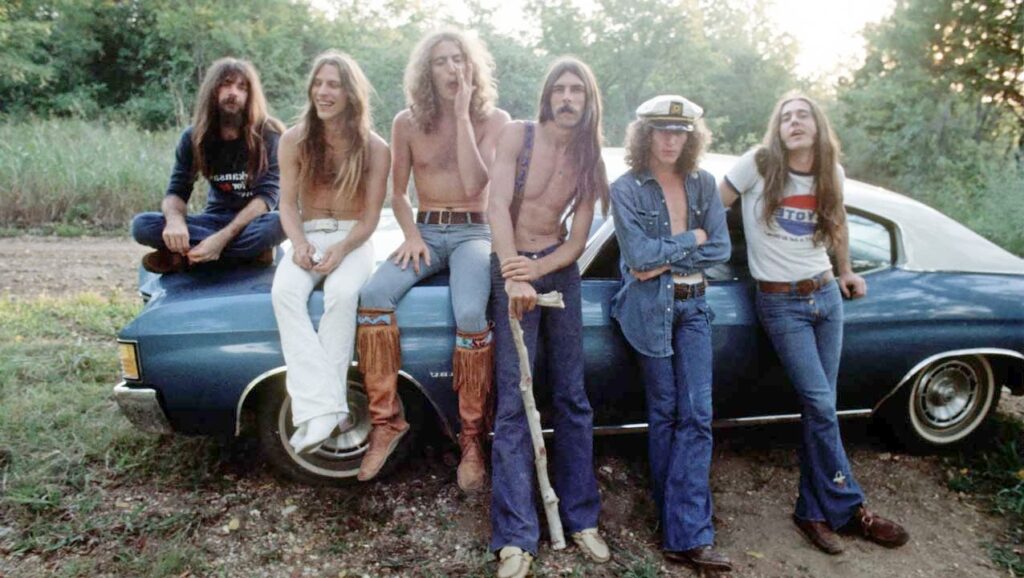 Brian Johnson (left) poses with his 1970s rock band Geordie in front of a classic muscle car, showcasing long hair, flared jeans, and vintage style during their early UK glam rock era.