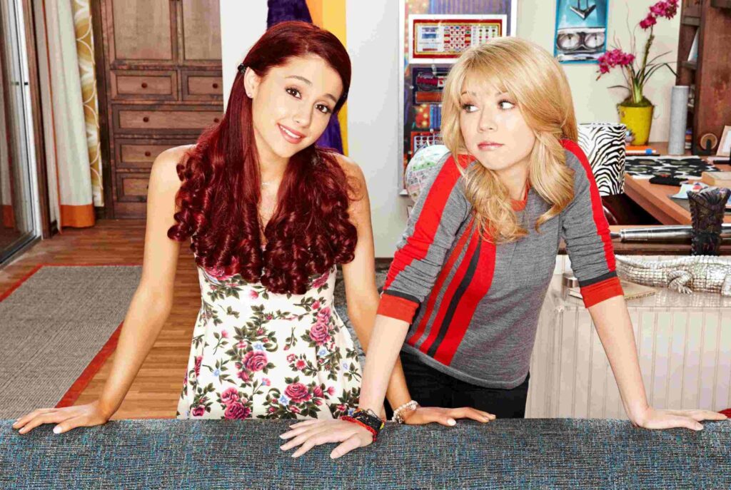 Ariana Grande with long red curly hair wearing a floral dress, standing beside Jennette McCurdy in a colorful room from the TV show Sam & Cat.