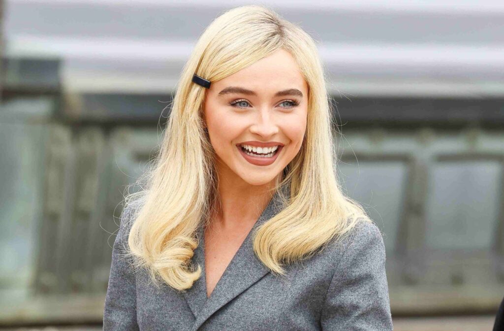 A smiling blonde woman in a grey outfit, posing outdoors, used as a style-inspired image for Sabrina Carpenter fashion content.