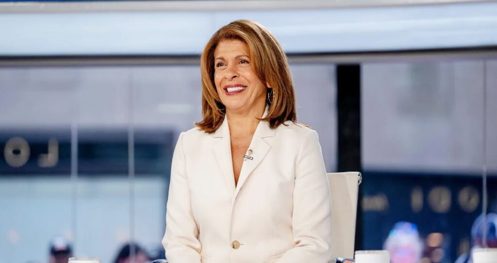 Hota Kodb wearing a light-colored blazer smiles while seated in a professional studio setting with blurred background visuals.”
