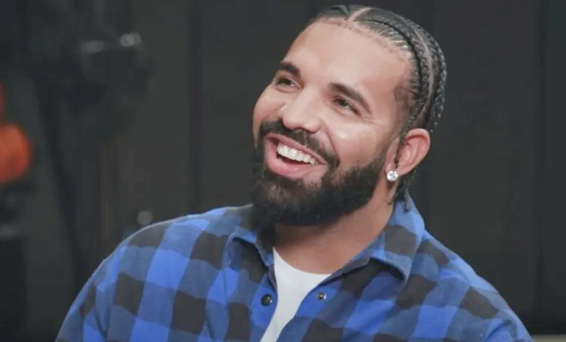"A man with neatly braided hair, a full beard, and earrings smiling while wearing a blue and black checkered shirt."