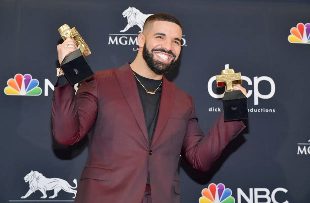 A smiling man in a maroon suit holding two golden music awards while posing in front of an event backdrop with NBC and MGM logos