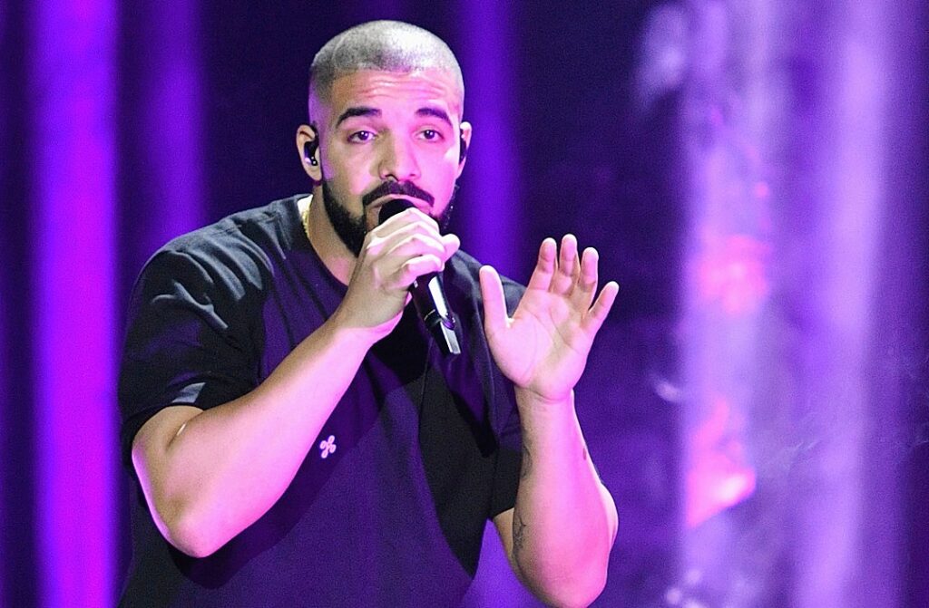 A male performer with a closely shaved haircut and beard sings into a microphone on stage, bathed in bright purple lighting while gesturing with his hand