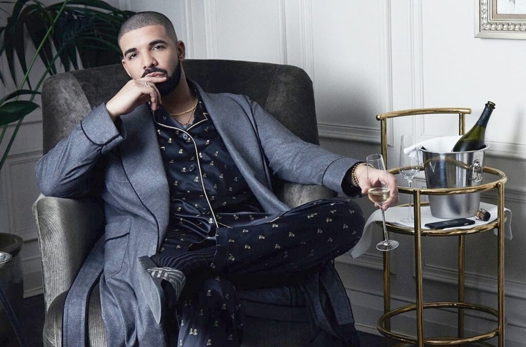 A well-dressed man sitting relaxed in an armchair, wearing a patterned pajama set with a grey blazer, holding a drink while resting beside a gold bar cart with a wine bottle in an ice bucket