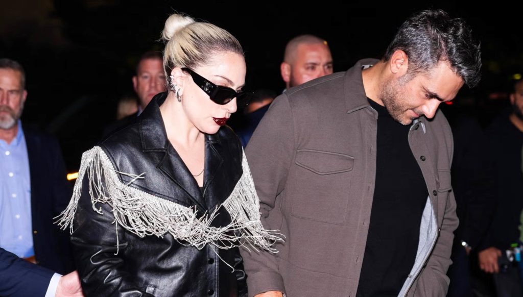 A woman in a black leather jacket with sparkling fringe walks beside a man at night, wearing dark sunglasses and bold makeup — image used in content related to Lady Gaga net worth.