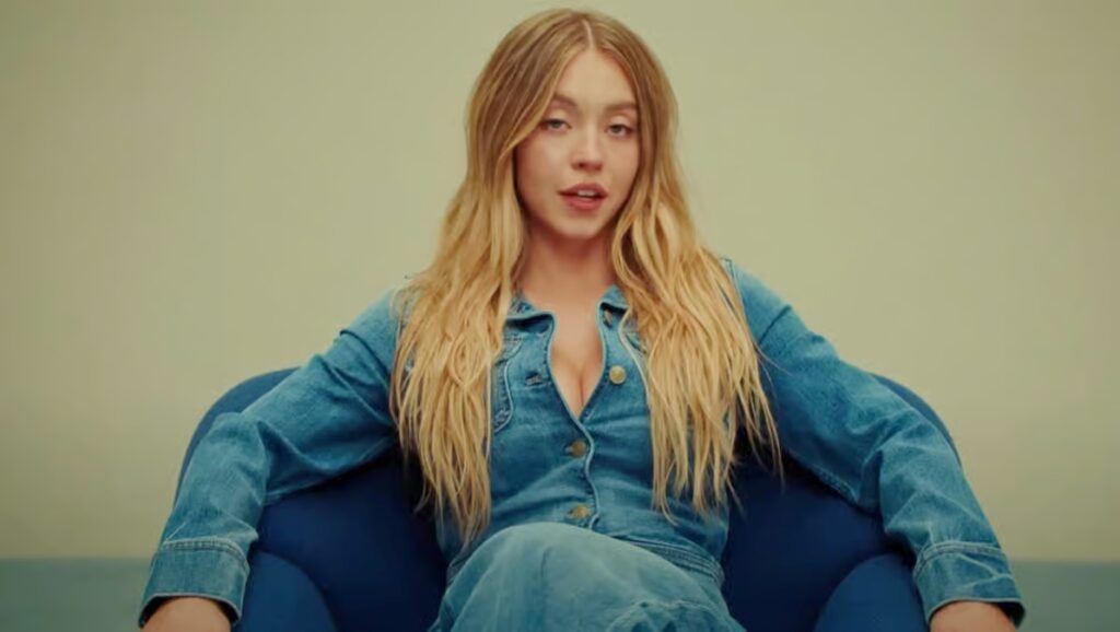 A woman with long blonde hair sitting against a plain background, dressed in a denim shirt and jeans, looking calmly toward the camera.