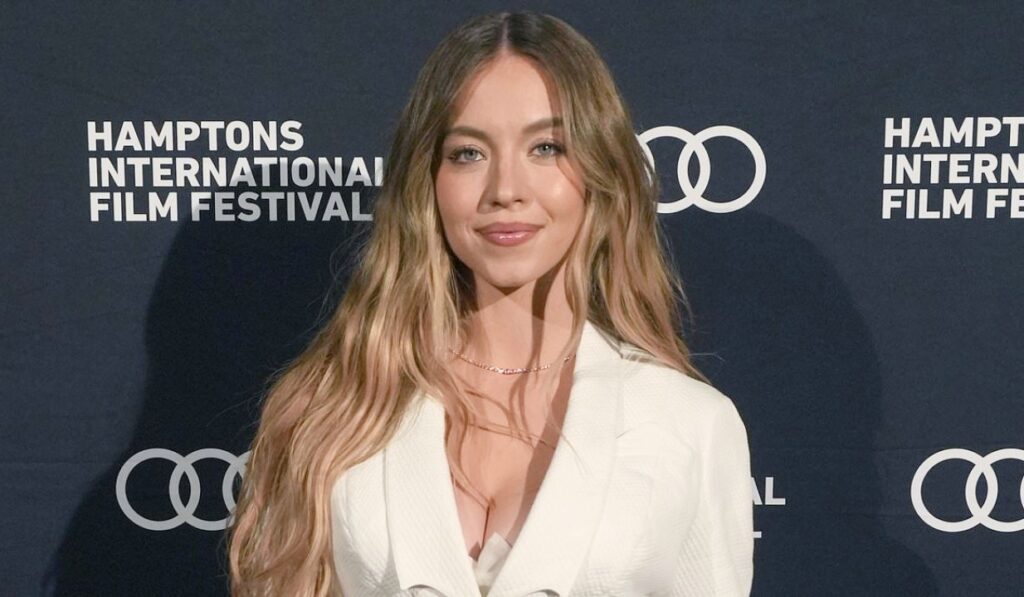 Woman with long wavy blonde hair wearing a white outfit, posing in front of a Hamptons International Film Festival backdrop.