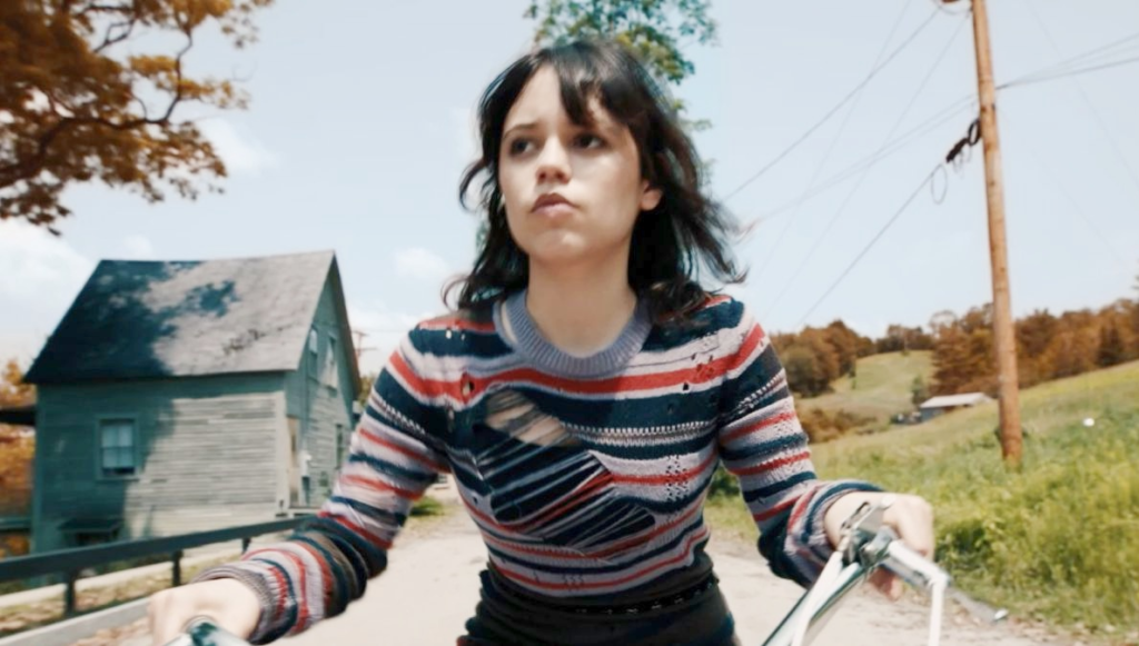 A young woman riding a bicycle along a rural road, wearing a striped sweater with a determined expression, suitable for articles related to Jenna Ortega net worth and celebrity lifestyle topics.