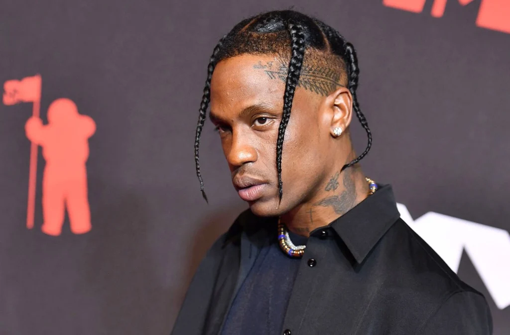 A close-up photo of Travis Scott at a red carpet event, showing his signature braided hairstyle, tattoos, and black outfit, suitable for articles discussing Travis Scott net worth and career highlights.