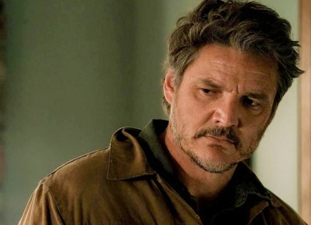 A rugged-looking man with tousled hair and a serious expression wearing a brown jacket indoors, featured in an article about Pedro Pascal Net Worth.