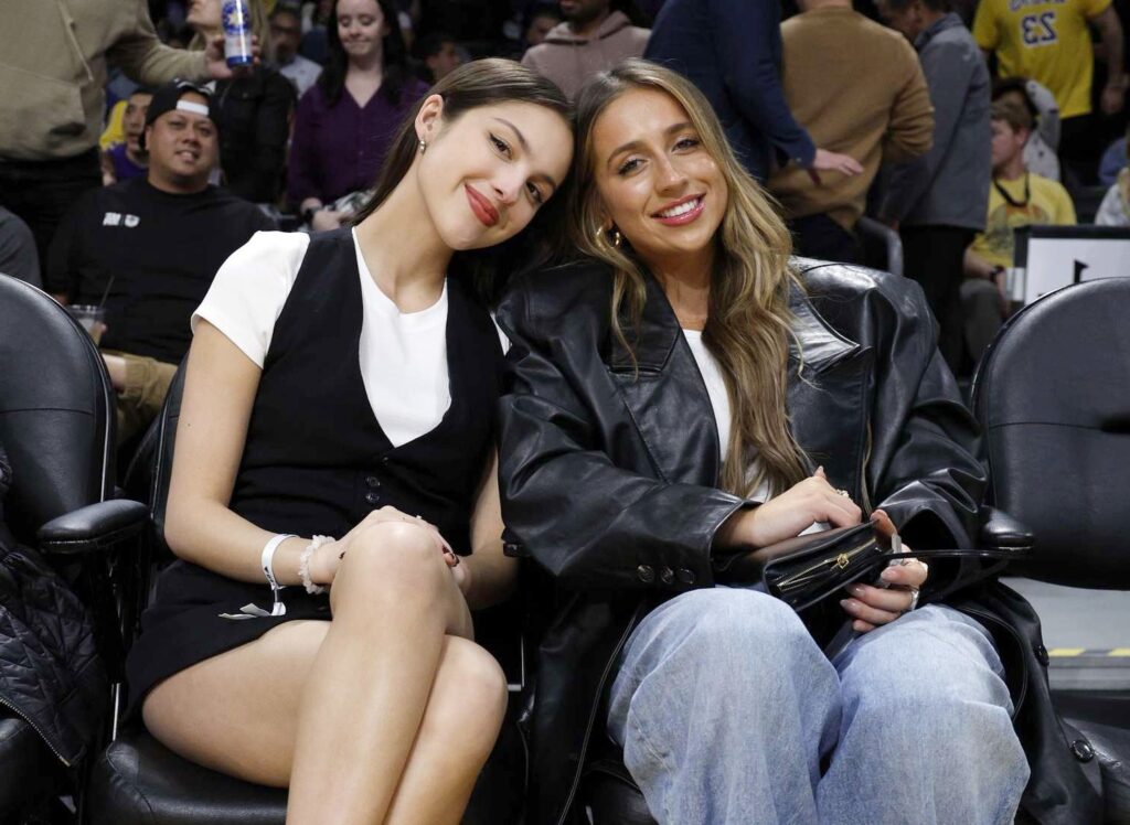 Two young women sitting courtside at a basketball game, smiling and enjoying the event, representing celebrity lifestyle and high-profile public appearances often linked with topics like Tate McRae net worth.
