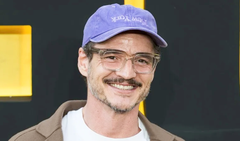 A smiling man wearing a purple cap, clear glasses, and a casual jacket, used to illustrate an article about Pedro Pascal Net Worth.