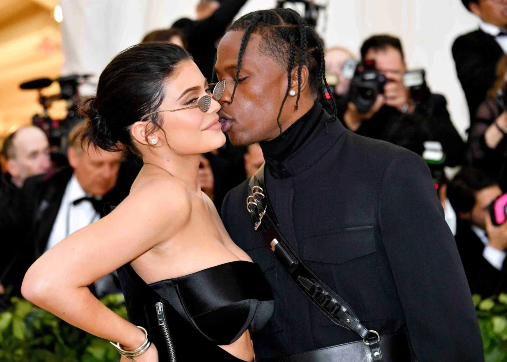 A stylish couple posing closely on a red carpet, surrounded by photographers, during a high-profile event—ideal for articles exploring Travis Scott net worth, celebrity relationships, and luxury lifestyle culture.