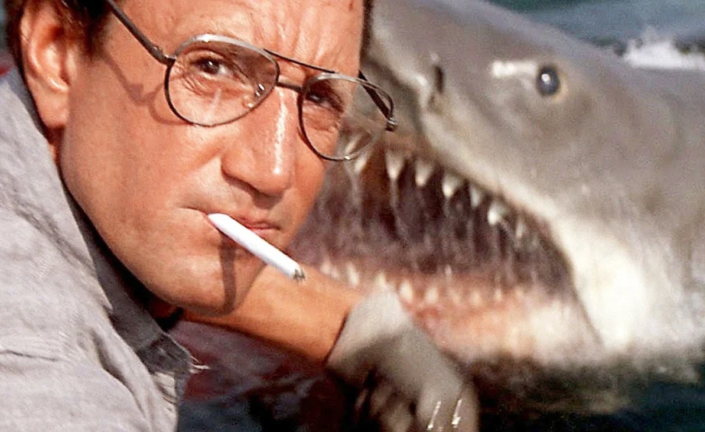 Steven Spielberg Net Worth related image showing a tense movie scene with a man wearing glasses and a cigarette, closely facing a giant shark.