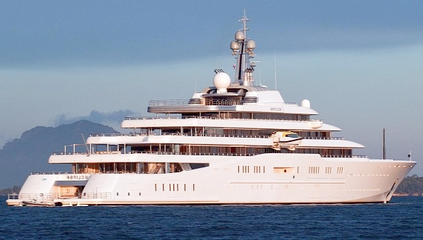 A luxurious multi-deck white megayacht sailing on the ocean with mountains in the background, featured in content discussing Roman Abramovich Net Worth.