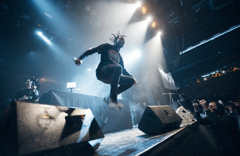 An energetic performer jumping mid-air on stage during a live concert, surrounded by dramatic lighting and photographers, suitable for articles discussing Travis Scott net worth and the impact of high-energy hip-hop performances.