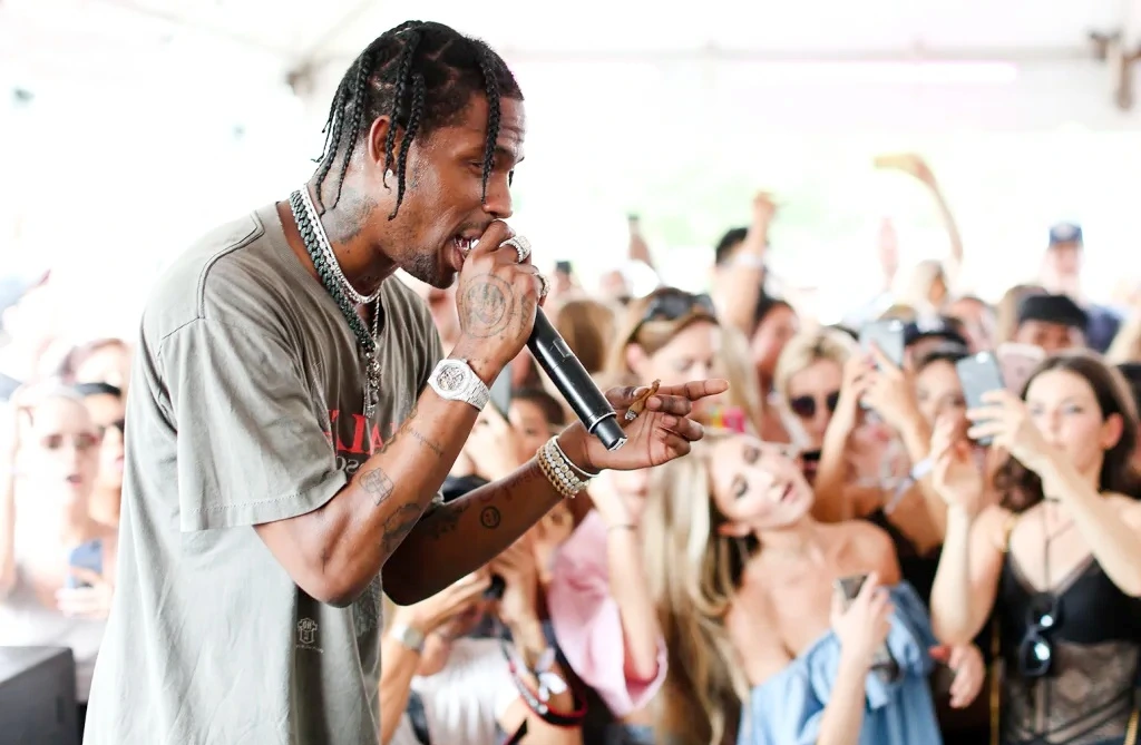 Travis Scott performing on stage in front of a large crowd, holding a microphone and wearing a casual T-shirt and jewelry, ideal for articles discussing Travis Scott net worth and his impact on music and live performances.